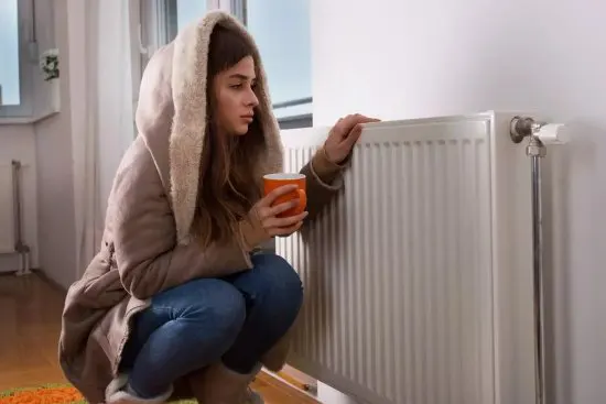 Girl in a coat leaning against a radiator due to a malfunctioning boiler caused by limescale.